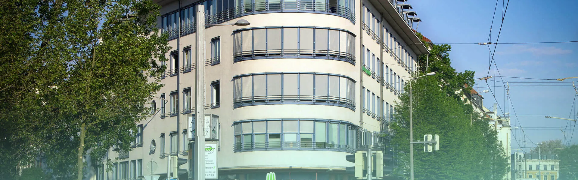 Modernes Eckgebäude mit geschwungener Glasfassade in einer begrünten Straße mit Straßenbahnlinien