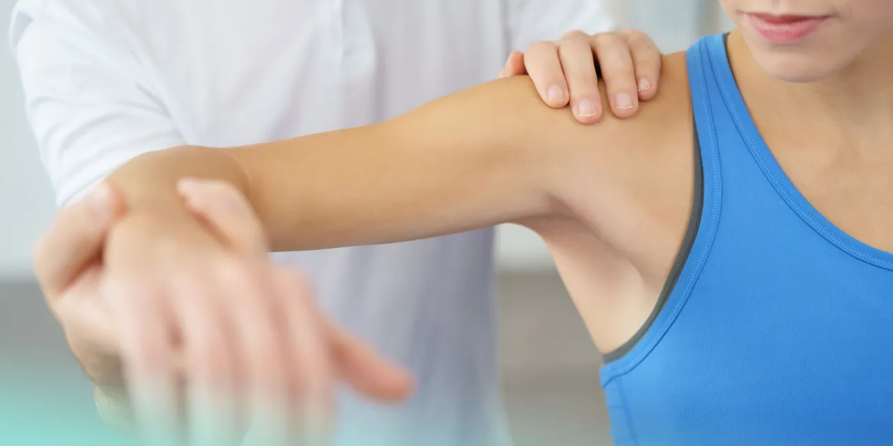 Physiotherapeut in weißem Kittel führt Armbehandlung bei Patientin in blauem Top durch