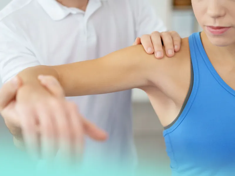 Physiotherapeut in weißem Kittel führt Armbehandlung bei Patientin in blauem Top durch