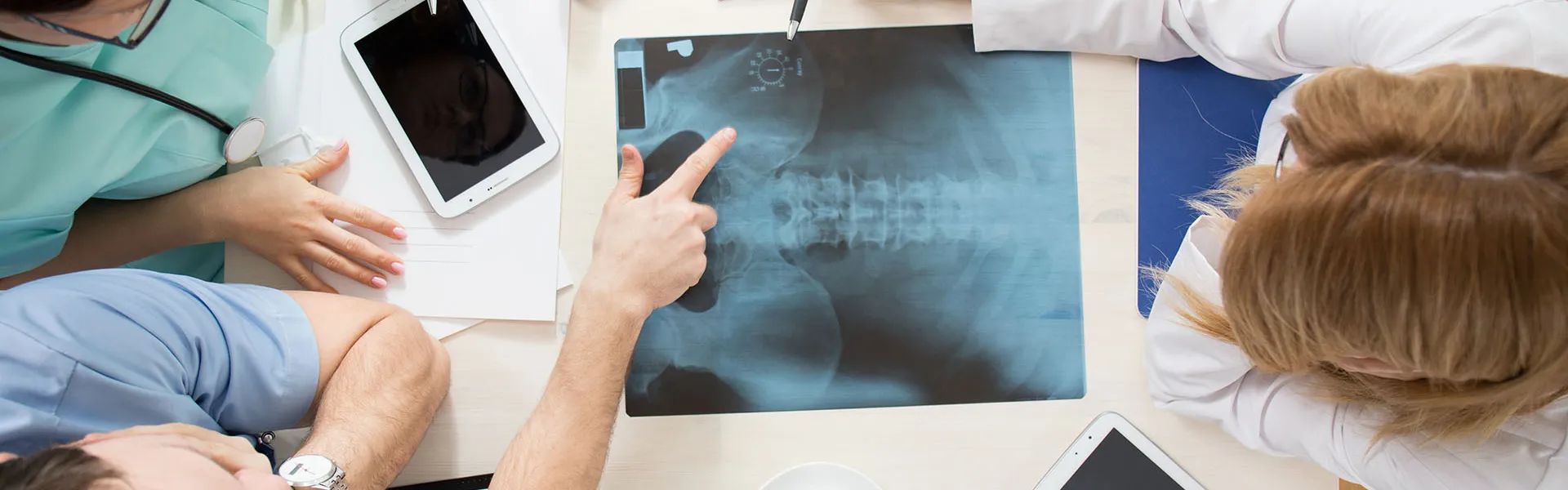 Ärzteteam bespricht Röntgenbilder am Tisch mit Tablets und Stethoskop in moderner Praxis