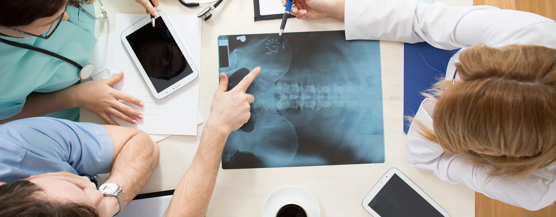 Ärzteteam bespricht Röntgenbilder am Tisch mit Tablets und Stethoskop in moderner Praxis